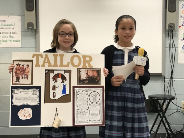 Two girls. One holds a poster that says Tailor, and the other holds a microphone.
