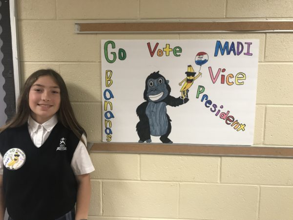 A girl stands in front of a school campaign poster that says Go Bananas Vote Madi Vice President